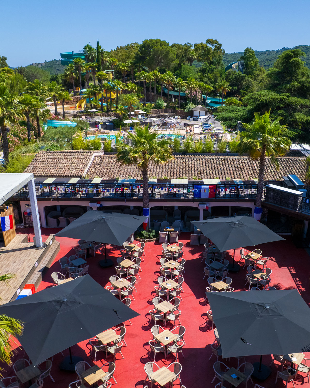 Terrasse du bar d'Esterel Caravaning sous ses parasols, avec les palmiers et les toboggans de l'espace aquatique en arrière-plan